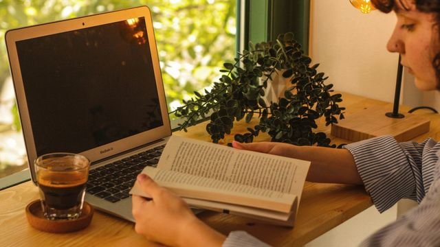 Laptop, livro e café numa mesa de madeira iluminada pelo sol