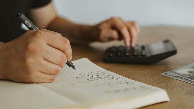 Mãos fazendo contas num caderno com calculadora